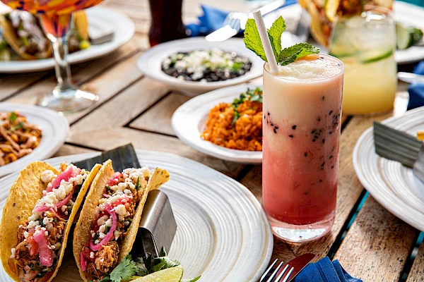 A lively outdoor meal: tacos, bowls, and dips on a wooden table with colorful drinks and garnishes, as friends dine al fresco.