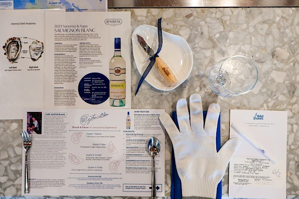 A stylish dining setup with a beer of white wine, a shelled seafood dish, a tortilla-topped baked dish, and a bowl of oysters, in a bright modern restaurant.