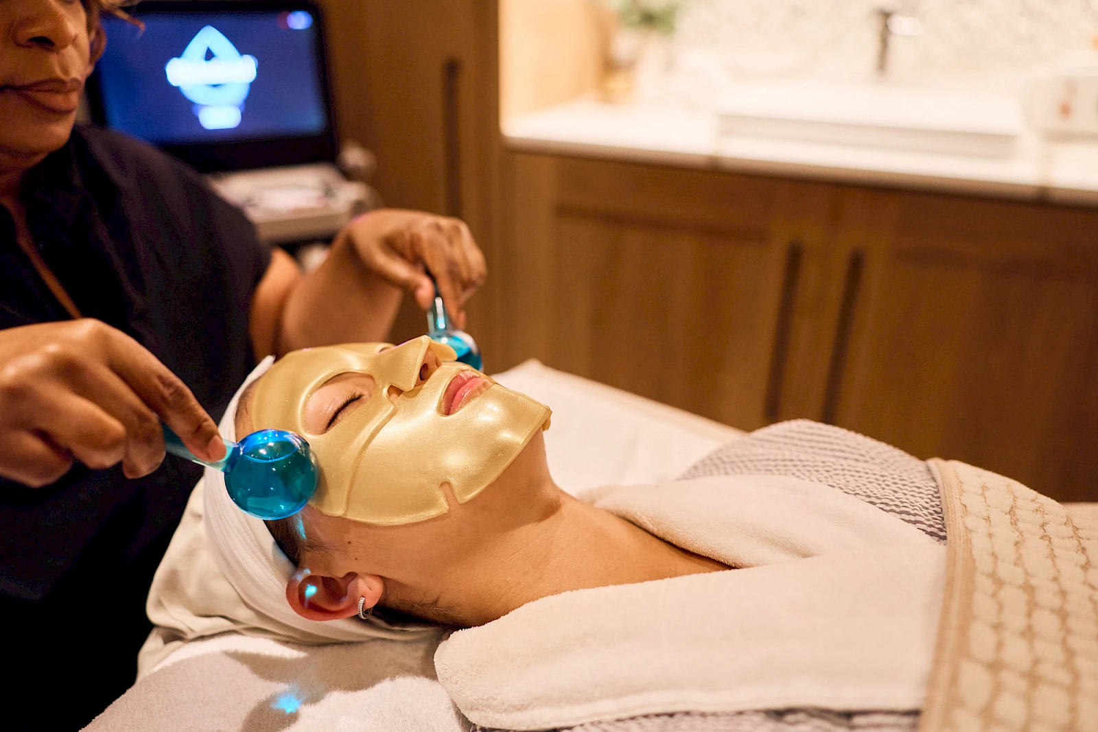 A person wearing a gold facial mask and eye protection receives a blue facial treatment with a nozzle and wand from a professional.