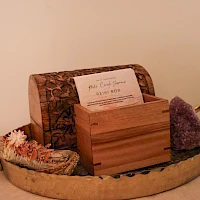 A round metal tray with two gold handles holds a wooden box, a carved wooden case, a bar of soap, and a purple crystal cluster on a beige background.