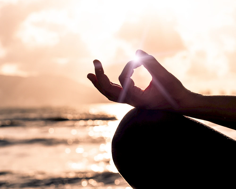 A pregnant person at the beach makes a hand gesture framing the sun, creating a serene sunset moment with soft waves in the background.
