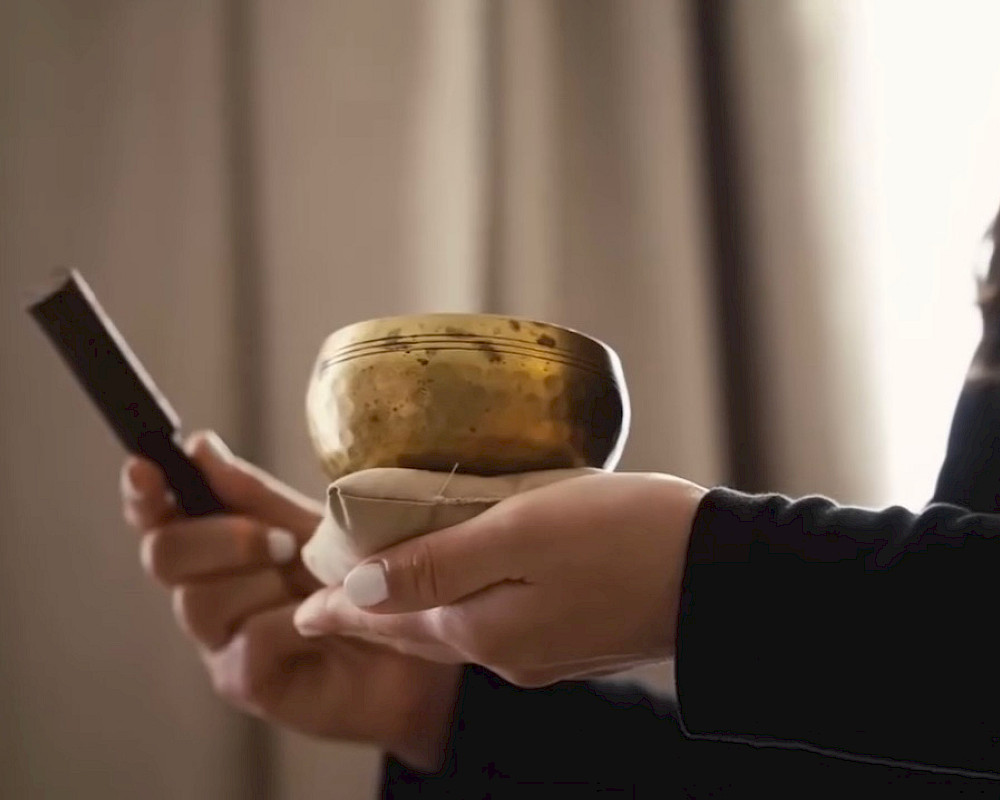A person holding a small brass singing bowl in one hand and a hammer or striker in the other, likely about to strike it.