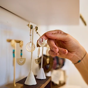 A hand holds a pair of dangling striped earrings among a display of various handmade jewelry on stands, in a shop. End with a period.