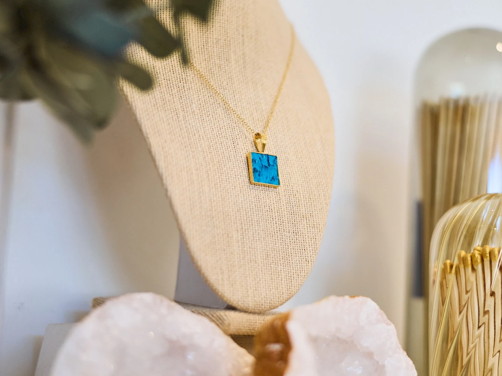 A decorative display with a turquoise square pendant necklace on a beige stand, flanked by crystals and geode slices, plus a glass holder in the background.