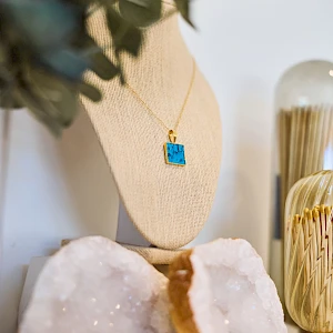 A decorative display with a turquoise square pendant necklace on a beige stand, flanked by crystals and geode slices, plus a glass holder in the background.