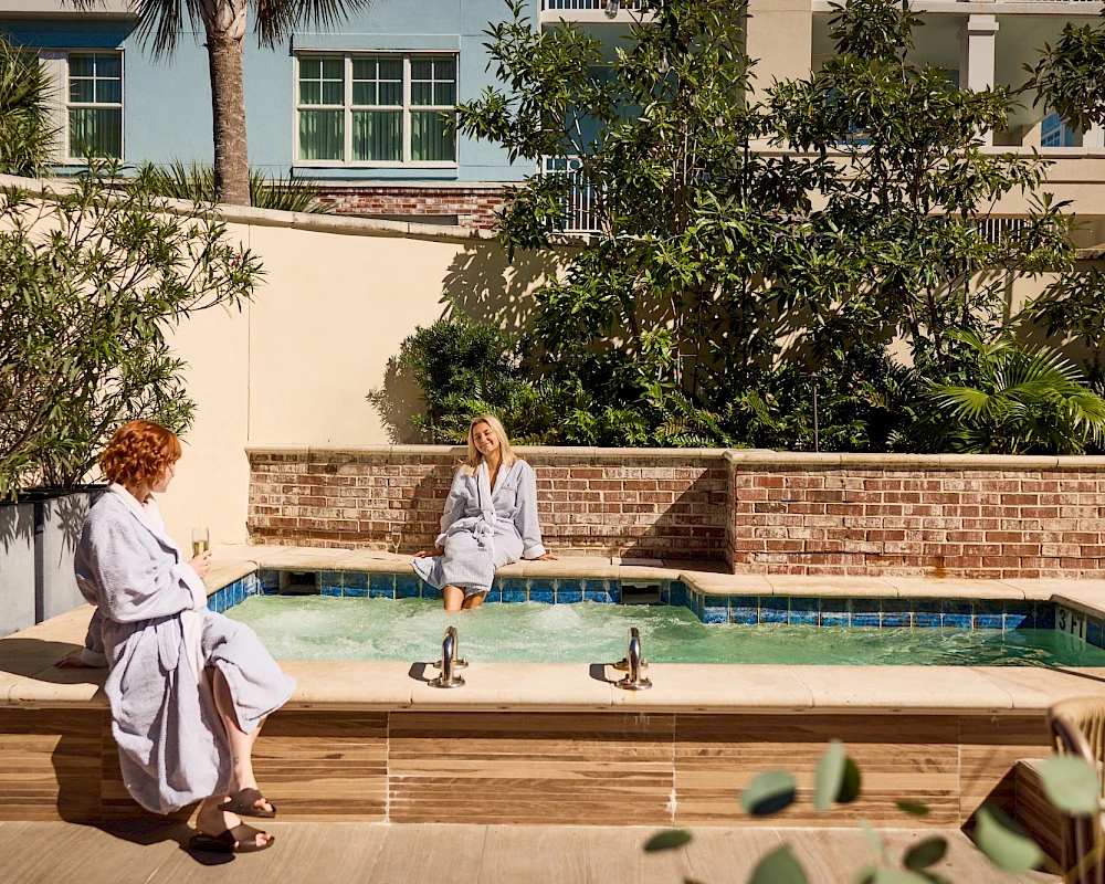 Two people sit by a shallow pool in robes, chatting beside a brick wall and lush green trees near a sunny courtyard.