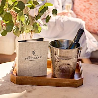 A small wooden tray with a plant and a metal ice bucket sits on a table; a cozy outdoor scene with soft cushions in the background.
