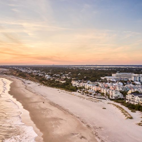 Aerial view of a beach at sunset with ocean waves, sandy shore, and a coastal town with colorful houses and greenery.