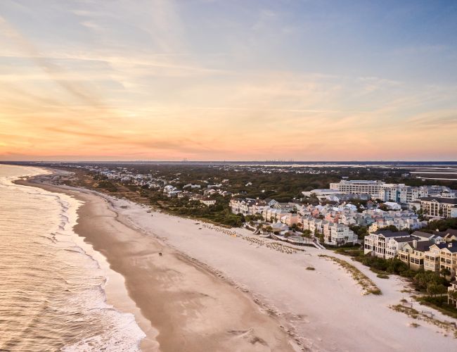 Aerial view of a beach at sunset with ocean waves, sandy shore, and a coastal town with colorful houses and greenery.