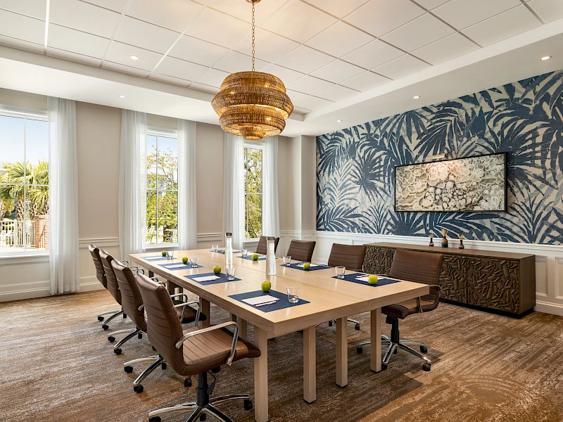 A modern conference room with a long wooden table, brown chairs, blue tropical wallpaper, windows with white curtains, and a woven pendant light, meeting setup complete.