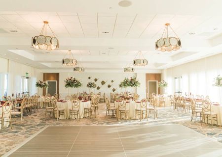 An elegantly decorated banquet hall features round tables with floral centerpieces, surrounded by chairs under large modern chandeliers.