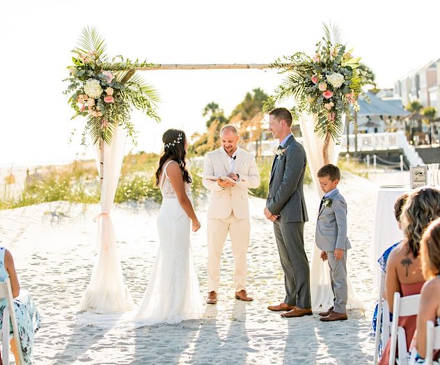 A beach wedding ceremony with a couple, officiant, and guests, under a floral arch on a sunny day.
