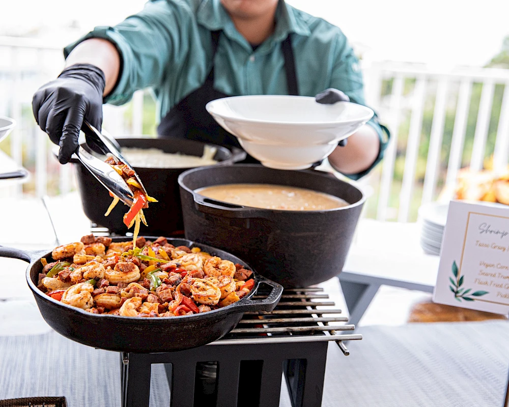 A person grilling food on a barbecue, tongs in hand adding pieces to a frying pan; another pan with sauce nearby, with a person holding a plate.