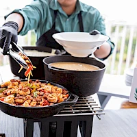 A person grilling food on a barbecue, tongs in hand adding pieces to a frying pan; another pan with sauce nearby, with a person holding a plate.