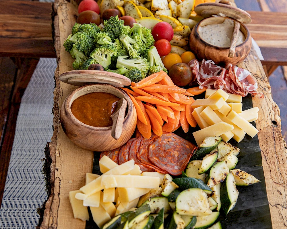 A long charcuterie board with assorted cheeses, sliced meats, olives, cherry tomatoes, crackers, roasted veggies, dips, and fresh herbs on a rustic wooden board.