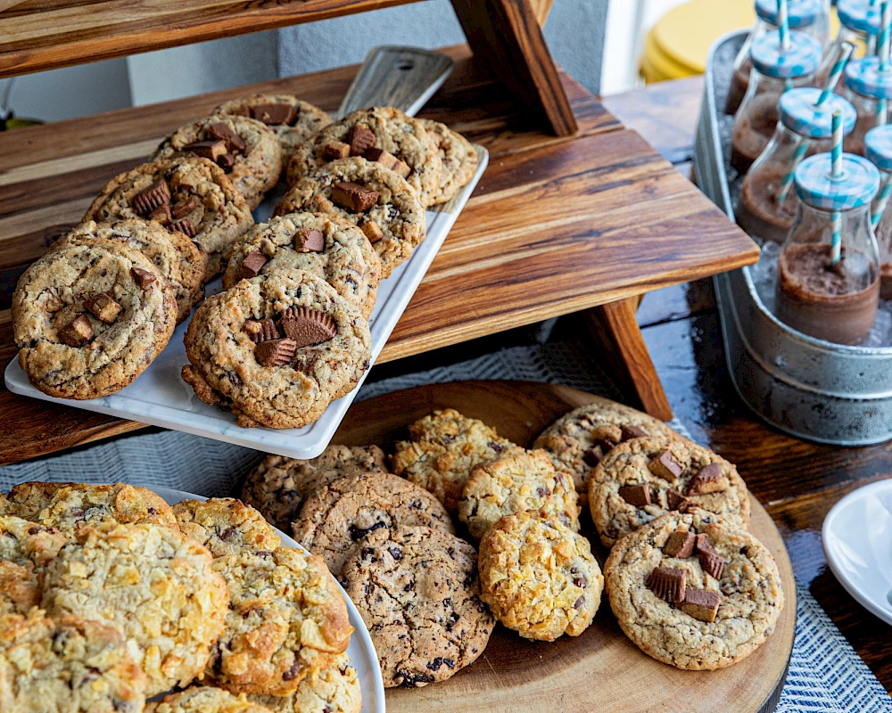 A display of assorted cookies on a wooden rack and tray, with nutty, chocolate chips, and crumbly textures, plus slim jars in the background.