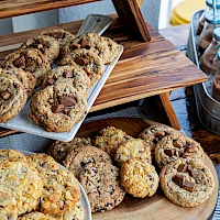 A display of assorted cookies on a wooden rack and tray, with nutty, chocolate chips, and crumbly textures, plus slim jars in the background.