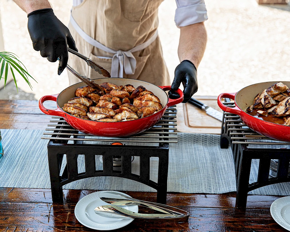 Two chefs grill skillet dishes outdoors on small portable grills, flipping meat to serve sizzling bites beside plates and cutlery.
