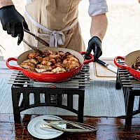 Two chefs grill skillet dishes outdoors on small portable grills, flipping meat to serve sizzling bites beside plates and cutlery.