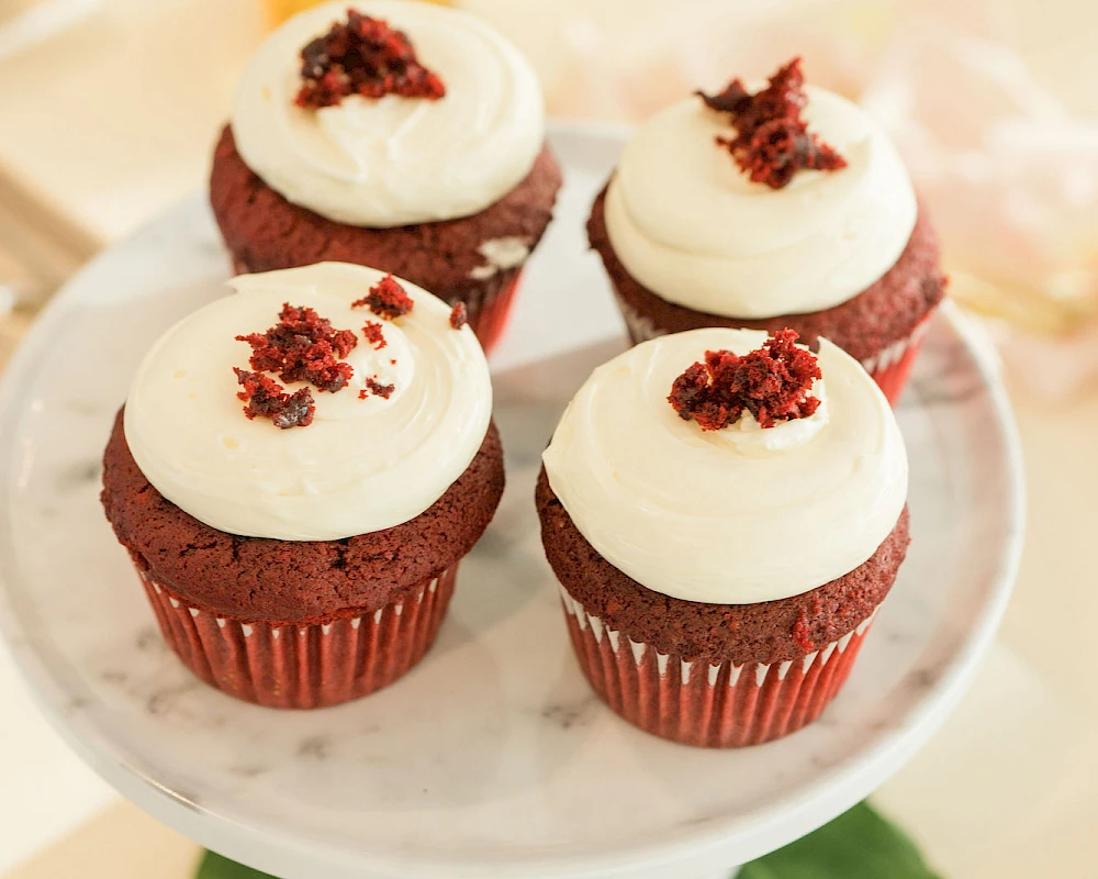 A dozen cupcakes with white frosting topped with red crumb bits, on a cake stand with a glass and flowers in the background.