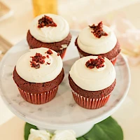 A dozen cupcakes with white frosting topped with red crumb bits, on a cake stand with a glass and flowers in the background.