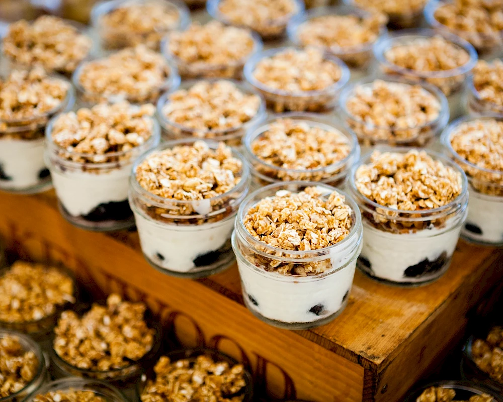 A countertop display of layered dessert cups with cream, cookies, and crunchy granola topping in small glass jars, arranged neatly.