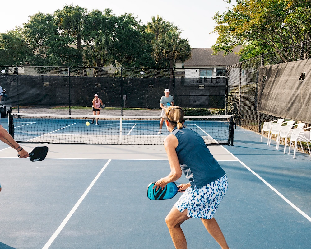 Three people play pickleball on a sunny outdoor court while others watch; one player swings at the ball as another prepares to return.