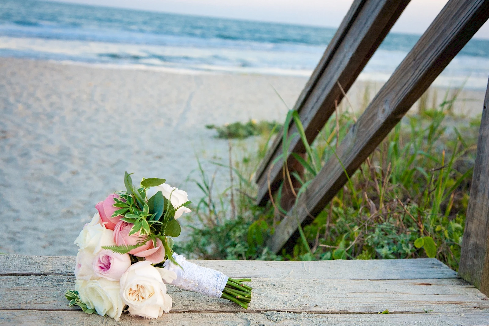 A small bridal bouquet with pink and white roses rests on a wooden deck by the beach, with dunes and sea in the background.