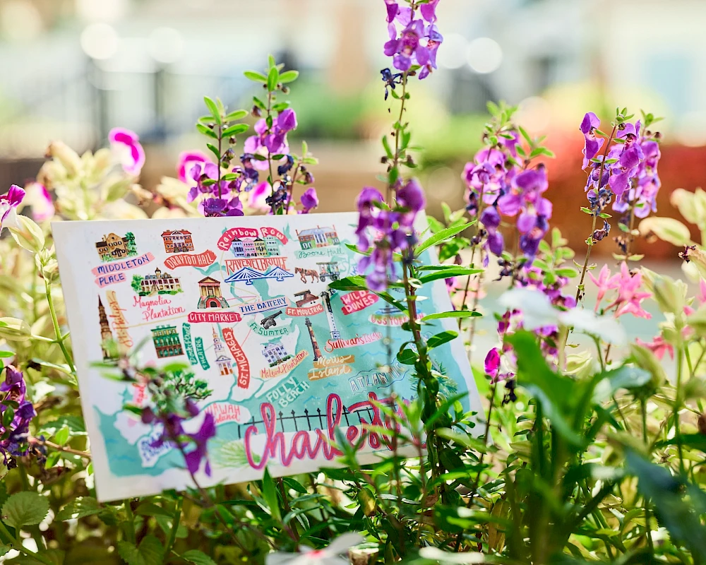 Colorful garden with purple flowers and a cartoon postcard tucked among the greenery, bright and cheerful scene finishing with a flower-filled charm.