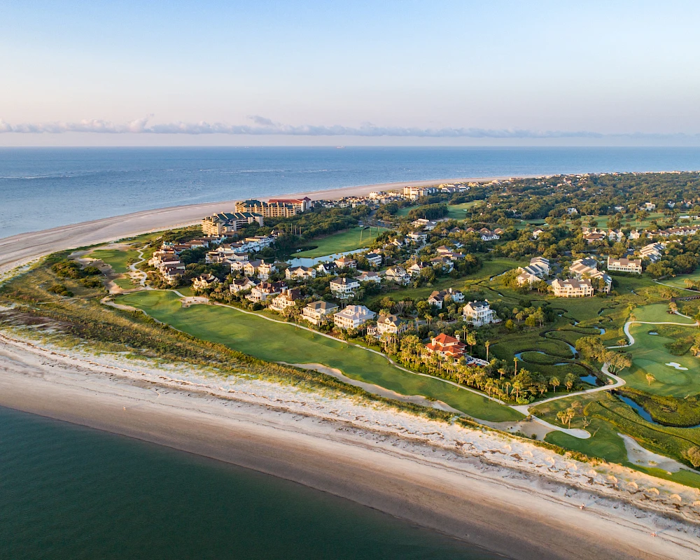Aerial view of a coastal residential area with sandy beach, green lawns, and homes along the shoreline, surrounded by water and blue skies.