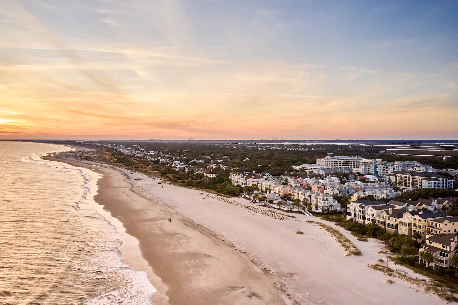 A coastal sunset over a sandy beach with calm waves and a cluster of white buildings lining the shore, bathed in warm orange-pink light.