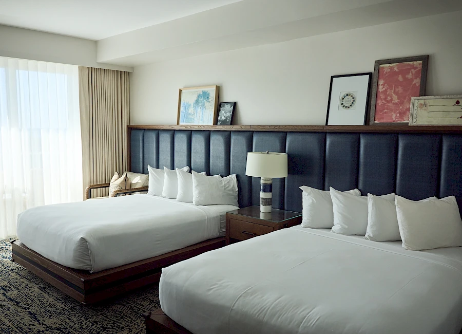 A hotel room with two large beds, a dark padded headboard, bedside tables, a lamp, and framed artwork, plus a window with sheer curtains.