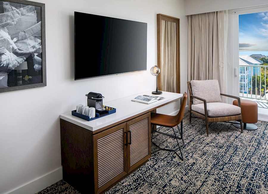 A stylish hotel room with a wall-mounted TV, a desk with a lamp, a small cabinet, a coffee/tea station, and a seating area by the window railing.