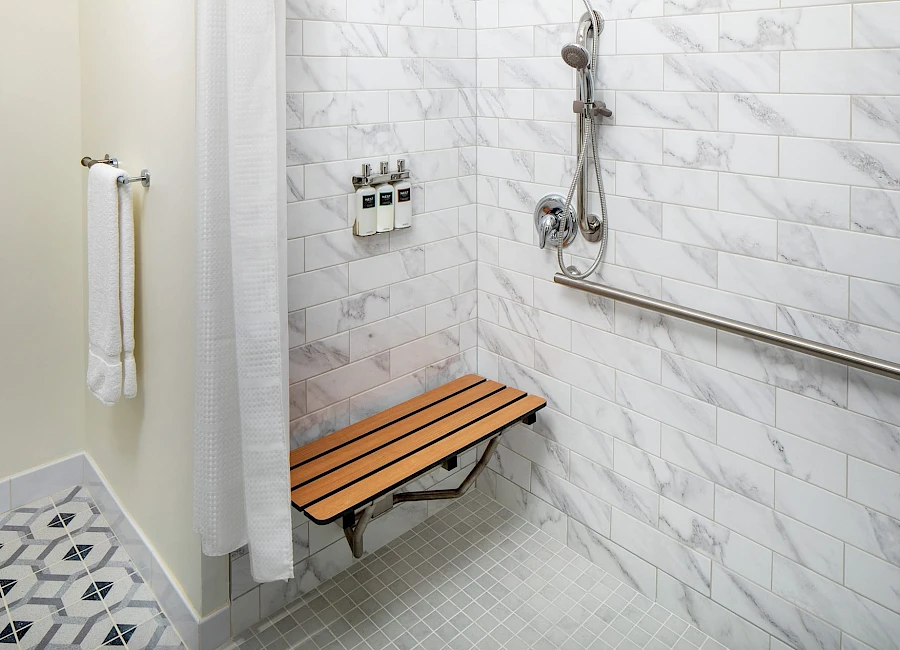 A modern accessible bathroom with marble walls, a wooden folding bench, a handheld shower, a vertical grab bar, and a towel rack beside a shower curtain.