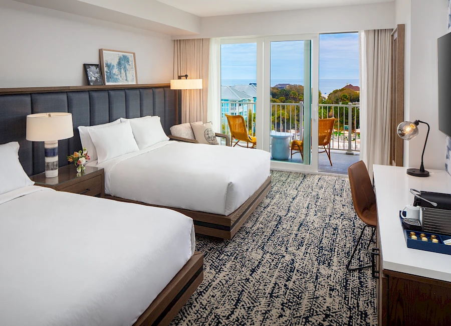 Two neatly made hotel beds in a bright room, with a desk, chairs, and a sliding glass door leading to a balcony overlooking a view.