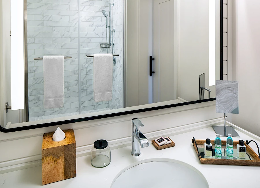 A modern bathroom sink area with a large backlit mirror, marble-tiled shower, towel racks, tissue box, glass jar, and a wooden tray of toiletries.