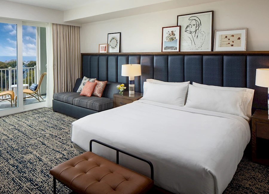 A modern hotel room with a large bed, blue padded headboard, nightstands with lamps, a dark sectional sofa, art prints above the bed, a brown bench at the foot, and a balcony view of mountains and blue sky.