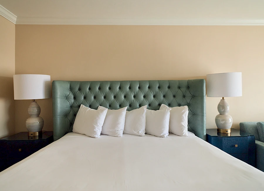 A neatly made bed with a tufted teal headboard, white linens, and several white pillows, flanked by matching nightstands and lamps.