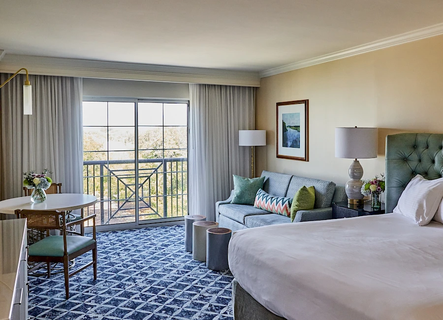 A hotel room with a king bed, gray upholstered headboard, sofa, round table with chairs, and a balcony view through glass doors, bright and airy.