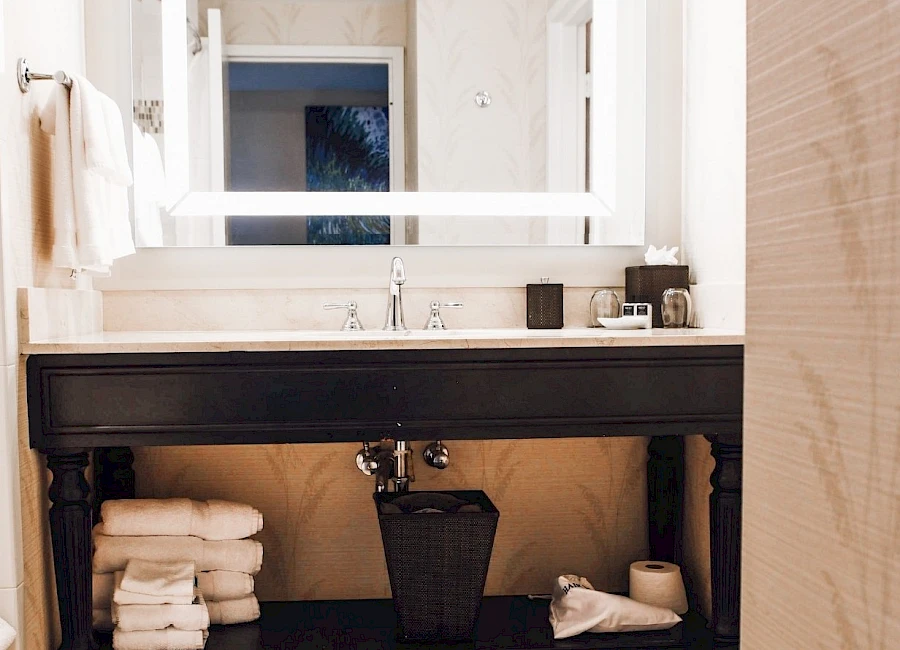 A modern bathroom with a black vanity, a large illuminated mirror, stacked towels on the shelf, and a small basket and trash can under the sink.
