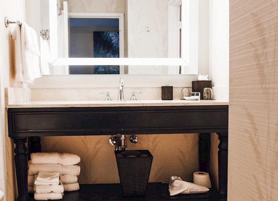 A modern bathroom with a dark wooden vanity, white illuminated mirror, stacked towels, a small trash bin, and a cozy, clean vibe.
