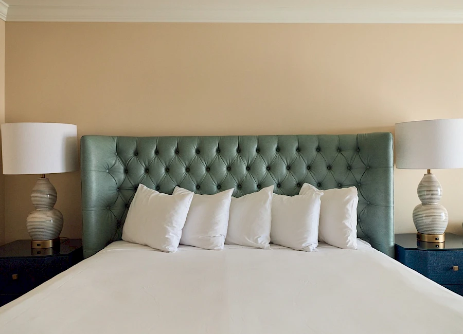 A neatly made bed with a tufted green headboard, five white pillows, matching nightstands, and two lamps flanking the bed.