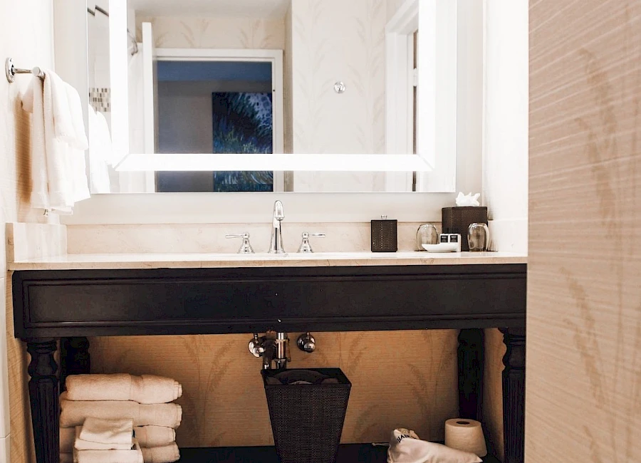 A modern bathroom vanity with a large backlit mirror, black wooden sink cabinet, folded towels, a white rug, and toiletries on the countertop.