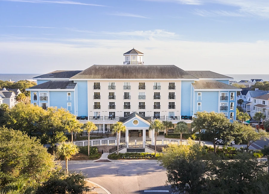 A large light-blue hotel or resort with a central cupola, rows of windows, a circular driveway, palm trees, and neatly landscaped grounds along a sunny coastal setting.