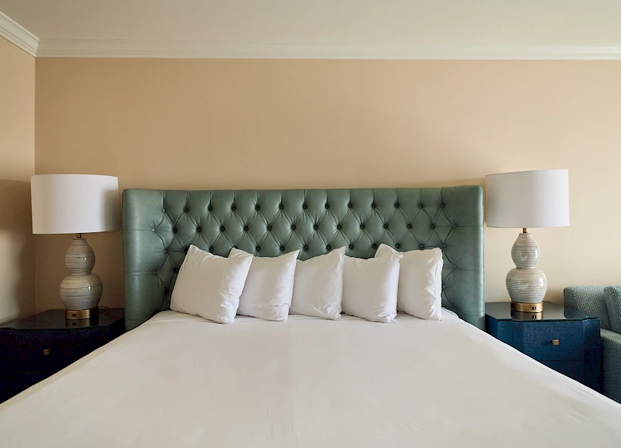 A neatly made bed with a tufted teal headboard, white linens, five white pillows, and matching bedside tables with lamps on each side.
