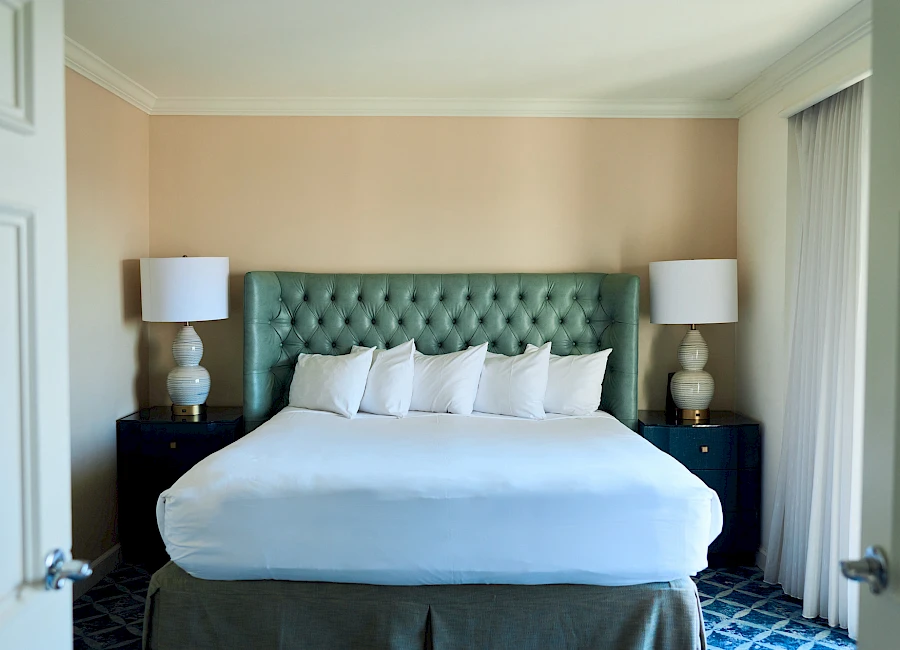 A neatly made hotel-style bedroom with a tufted green headboard, white bedding, two matching nightstands, and symmetrical lamps on each side.