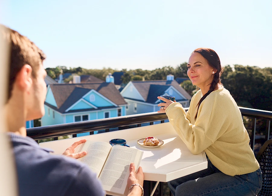 Two people chat on a balcony over coffee and snacks; a book lies on the table as they enjoy a sunny outdoor moment.