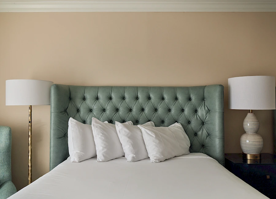 A neatly made bed with a tufted green headboard, white sheets, and multiple white pillows, flanked by two bedside lamps.