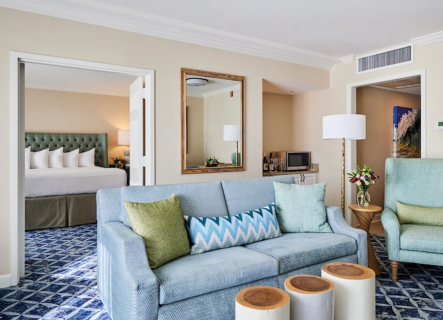 A bright hotel suite with a blue sofa and armchairs, patterned carpet, doorway to a bedroom, mirror, lamp, and a table with decor.