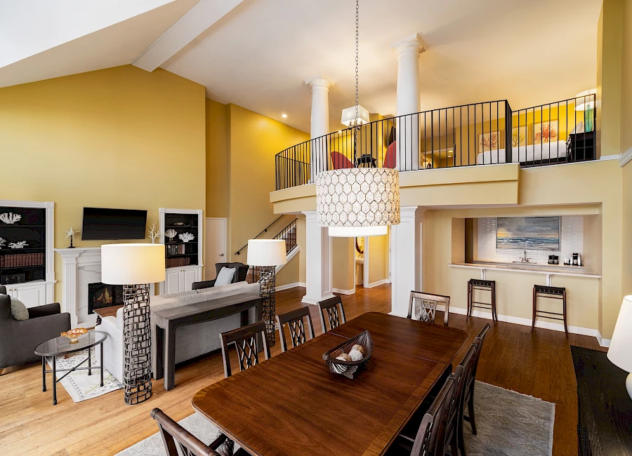 A spacious, modern living/dining area with a long wooden table, bright yellow walls, high ceiling, mezzanine balcony, and contemporary seating.